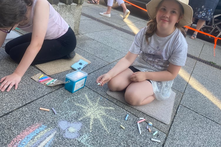 Young artists decorating Tavistock high street for the Lions Carnival Week pavement art competition. A girl chalks a flower, sun and rainbow for the theme 'golden moments'.