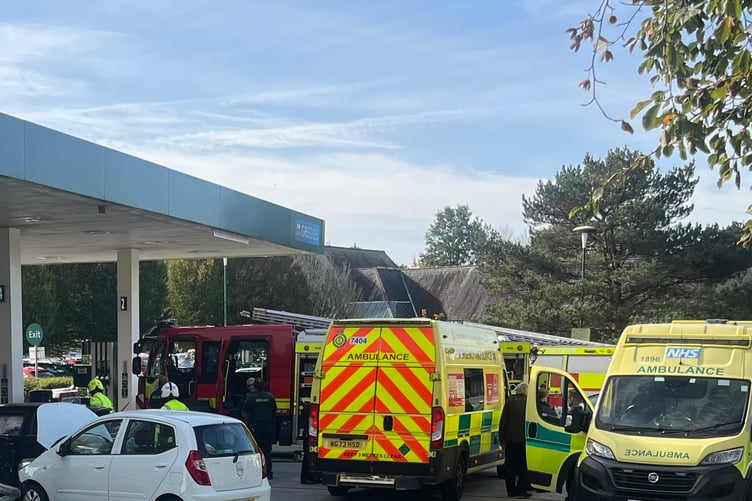 Fire engines, ambulances and police cars attended a car crash at Morrisons fuel station in Tavistock. Fire fighters in white hard hats and hi-viz tops are pictured to the left around a white car with its bonnet up.